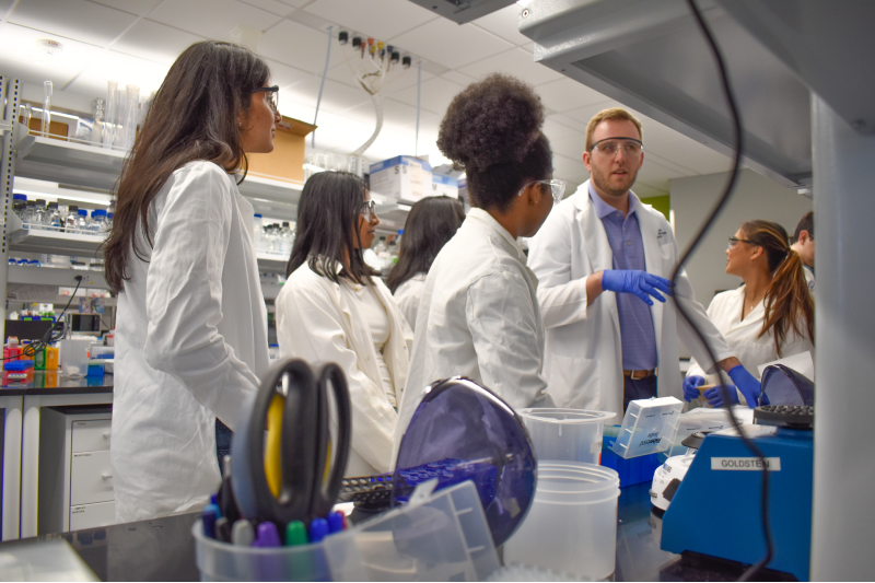 A male college professor speaks with a group of college students in a scientific lab.