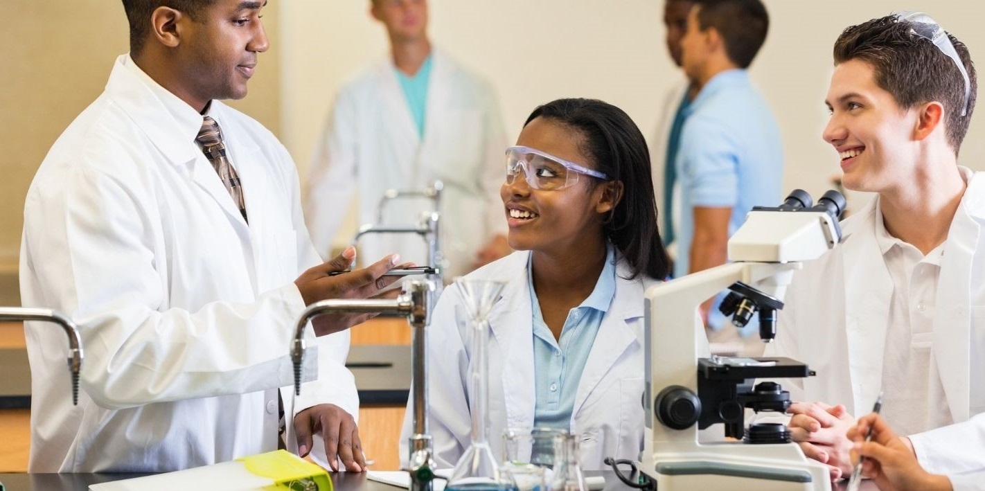Three young scientists in lab coats speak with each other in a lab.