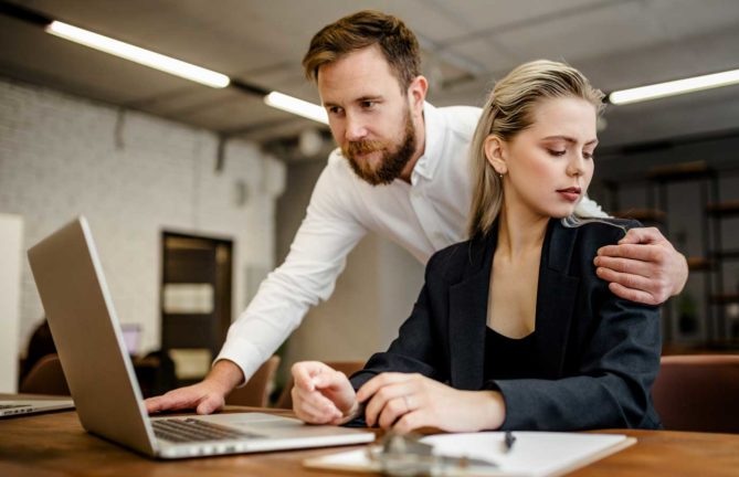 Man with arm around shoulder of woman looking at computer. Woman is uncomfortable with embrace.
