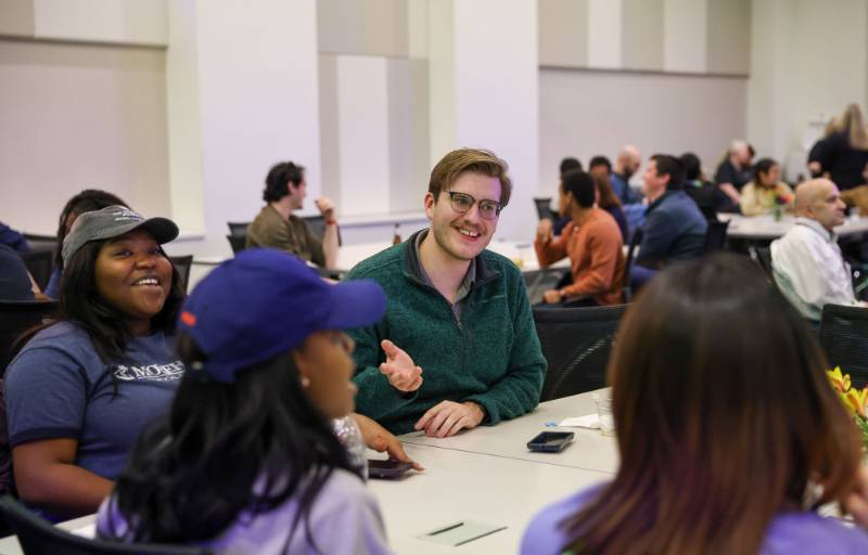 Students seated around a table smile and talk to one another