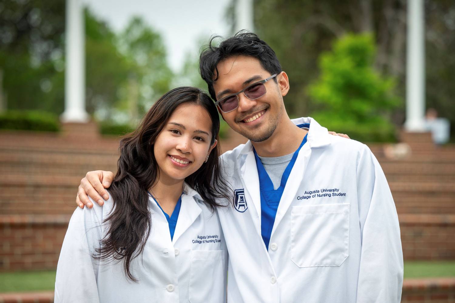 A man and woman both wearing white coats smile at the camera against an outdoor background with trees and bushes