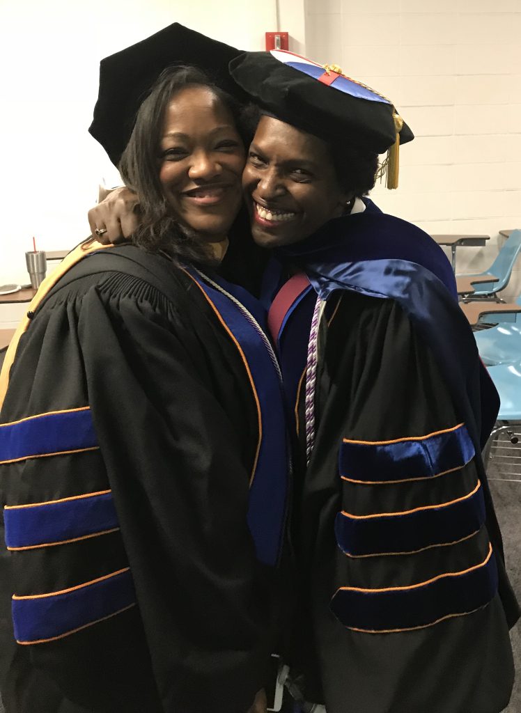 Two women in doctoral robes hug each other and smile at the camera