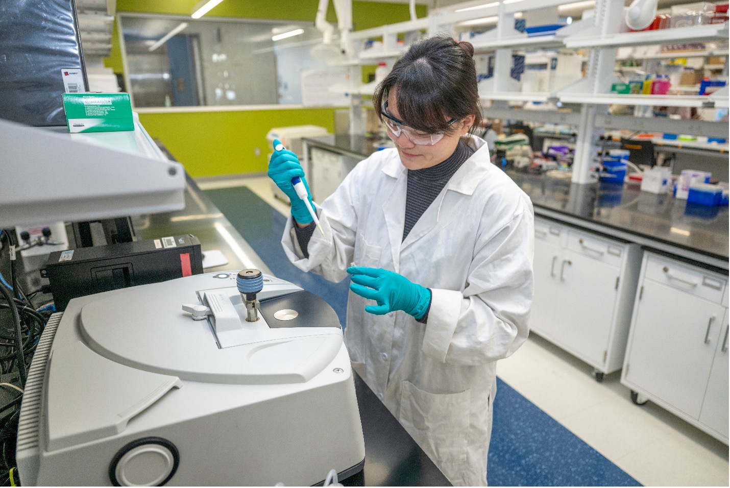 A woman in a white lab coat and safety glasses, Dr. Xiaobing Chen, pipettes in her lab. 