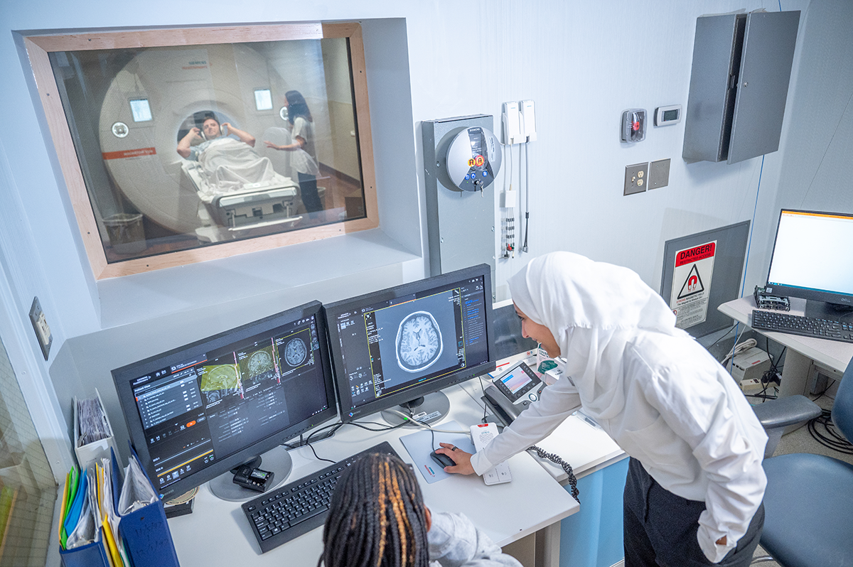 Two students look over the controls to an MRI machine, while another student is inside the machine for a photo