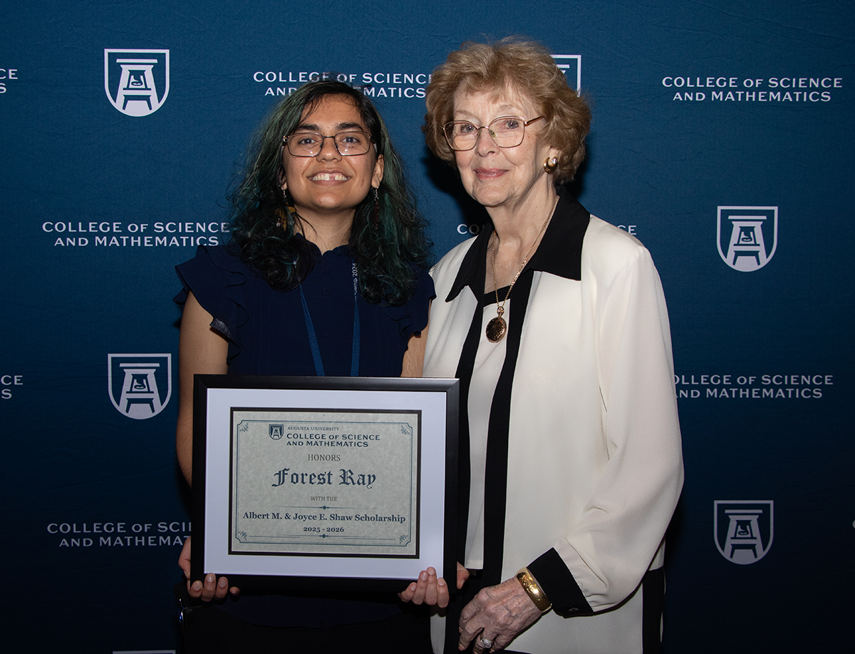Two women, (left: dark hair and blue dress; right: older, brown hair and off-white dress) stand holding an award at an honors night ceremony.