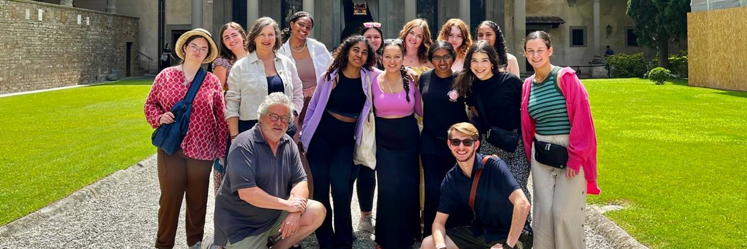 A group of people is posing outdoors in front of a historic building with columns and a dome. The setting is sunny with green grass on either side, and the group consists of individuals standing and two men kneeling at the front.