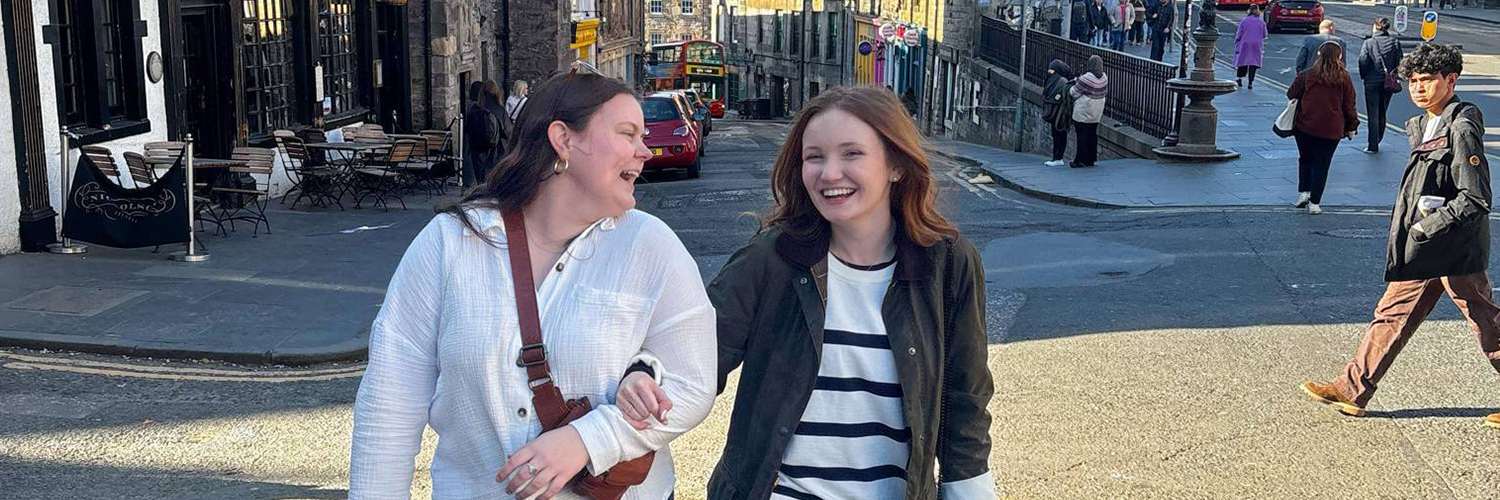 Two women walking arm-in-arm on a street in an historic urban area of Edinburgh, Scotland, with old stone buildings.