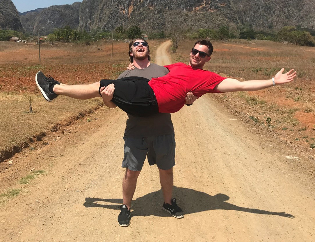 The image shows two people on a dirt road in a rural area with large rocky hills in the background; one person is standing and holding the other horizontally. The person being held is wearing a red shirt and black shorts, while the person holding them is dressed in gray. They both have wide smiles.