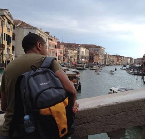 Brandon Dawson with a backpack on his back standing on a stone bridge overlooking a canal filled with boats in Venice, Italy. The background features historic buildings lining the waterway under an overcast sky.
