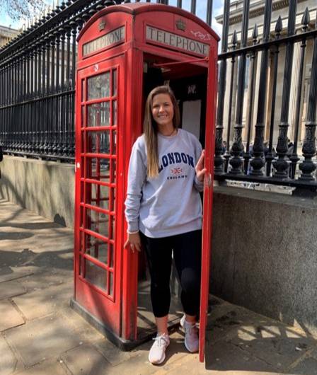 The image shows Brenna Johnson standing in the doorway of a classic red British telephone booth on a sidewalk next to an ornate black iron fence. She is wearing a light gray sweatshirt with “LONDON ENGLAND” printed on it, black leggings, and white sneakers, with sunlight casting shadows on the pavement.