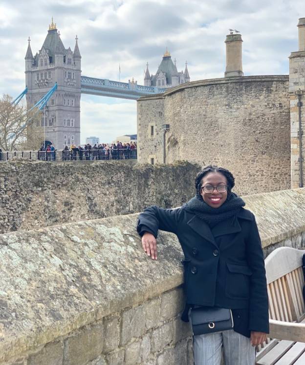 This image shows a person dressed in a dark coat and scarf, leaning against an old stone wall. In the background, the iconic Tower Bridge of London is visible under a partly cloudy sky, along with historic stone buildings that suggest the location is near the Tower of London. There are also several people gathered on a walkway behind the person, adding to the lively atmosphere of this popular tourist spot.