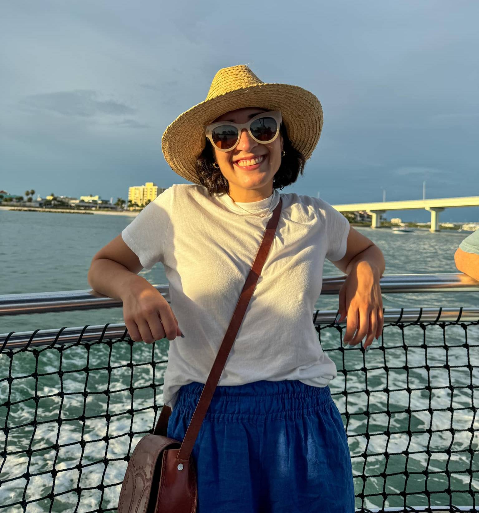 Elizabeth Canas wearing a straw hat, white t-shirt, and blue pants standing on a boat with water and a bridge in the background. She is leaning against the railing with both hands resting on it, and there is a brown crossbody bag hanging from her shoulder.
