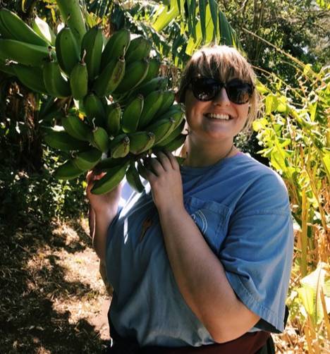 Kayla Hogan standing outdoors in a lush, green environment holding a large bunch of unripe bananas still attached to the plant. She is wearing a blue shirt and is surrounded by dense foliage and sunlight filtering through the leaves.