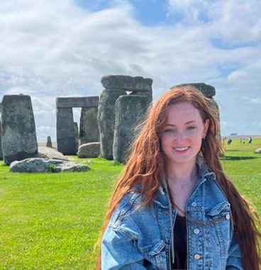 Neah Miles in a denim jacket stands near the Stonehenge monument on a grassy field under a partly cloudy sky.