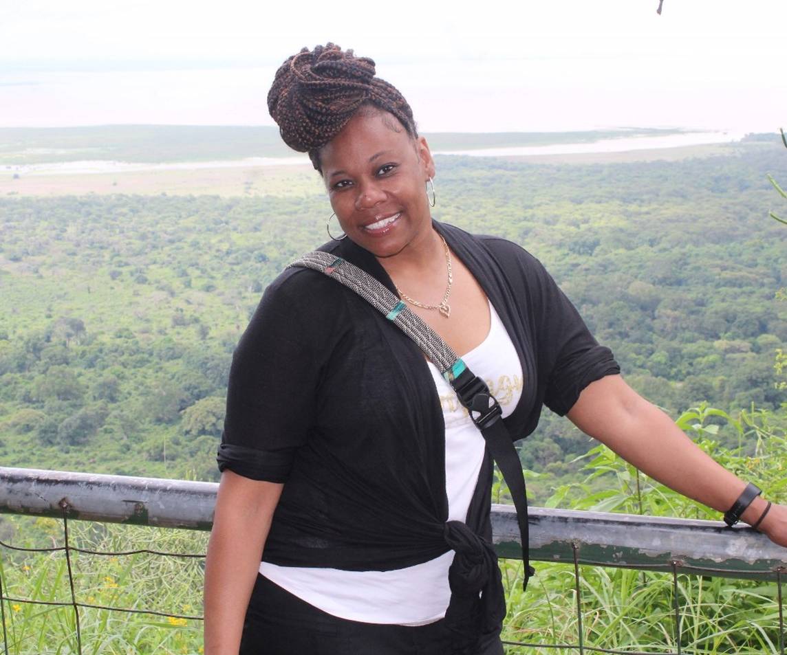 The image shows Sherita Cash standing outdoors, leaning on a metal railing with a vast green landscape and water body in the background. She is wearing a black cardigan over a white top, has braided hair styled up, and carries a crossbody bag strap across her chest.