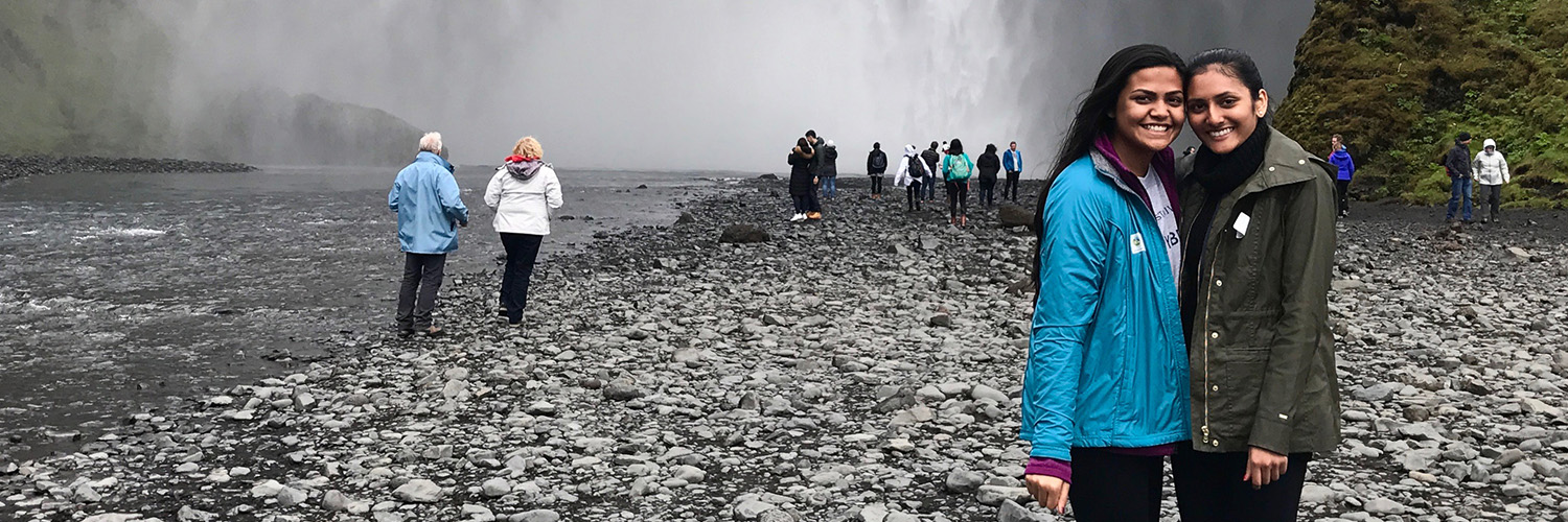The image shows a large waterfall with mist at its base, surrounded by rocky terrain and cliffs. Several people are walking or standing near the waterfall, including two individuals posing together in the foreground.