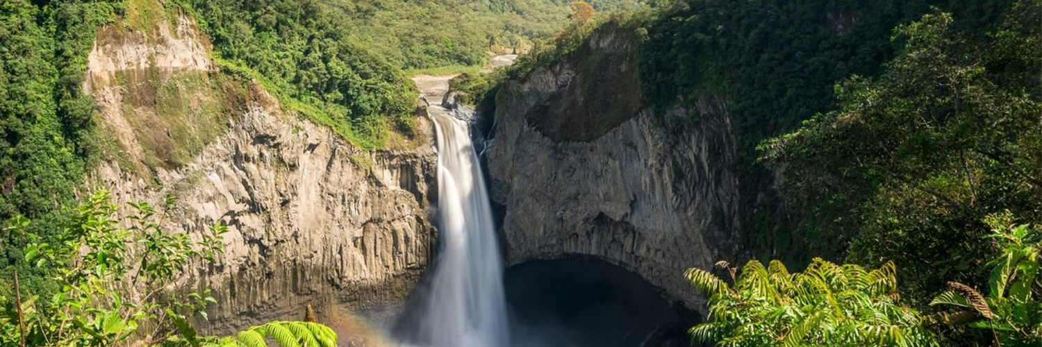 A tall waterfall plunging into a deep, circular gorge surrounded by dense, green forested mountains.