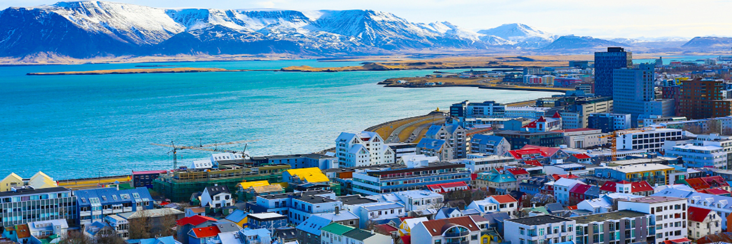 A vibrant coastal city with colorful buildings in the foreground and a large body of turquoise water extending towards snow-capped mountains in the background. The scene is bright and clear, highlighting both urban development and natural beauty under a partly cloudy sky.