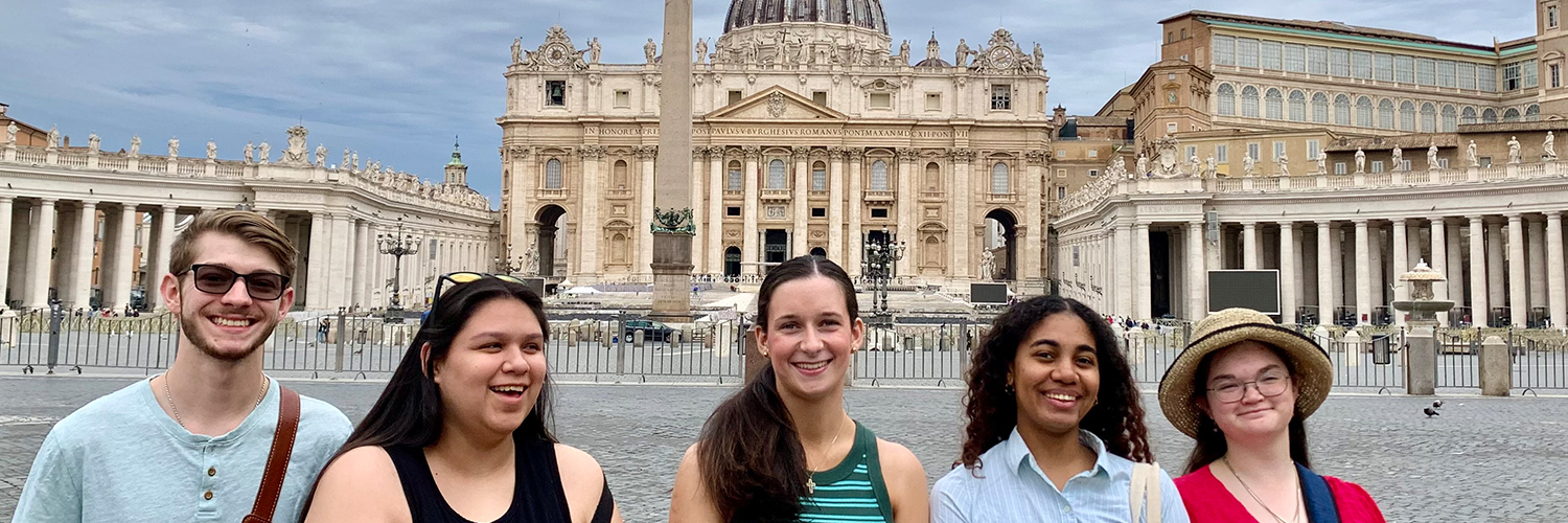 Group of five people standing in front of St. Peter’s Basilica in Vatican City, with the iconic dome and obelisk clearly visible behind them. The sky is overcast, and the people are posing for the photo on a cobblestone square.