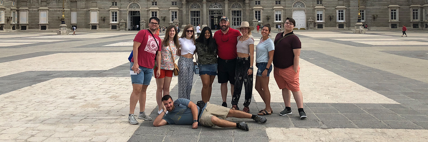 Nine people posing in front of the Royal Palace of Madrid, with one person lying down in front while the others stand behind. The large historic building features classical architecture and a Spanish flag flying on top under a partly cloudy sky.
